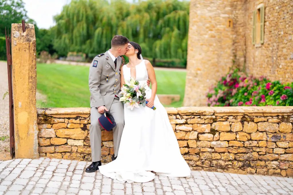 Mariés qui s'embrassent assis sur un muret en pierre près d'une tour, le marié en uniforme, la mariée avec son bouquet