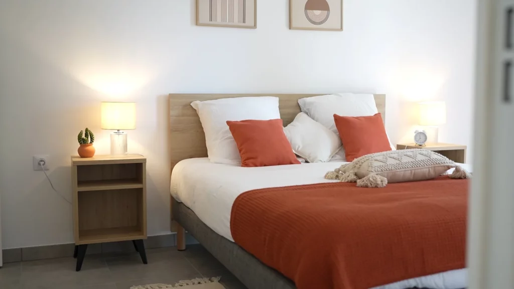 Chambre lumineuse et meublée d'un logement de la résidence, avec un lit aux coussins orange et deux lampes de chevet
