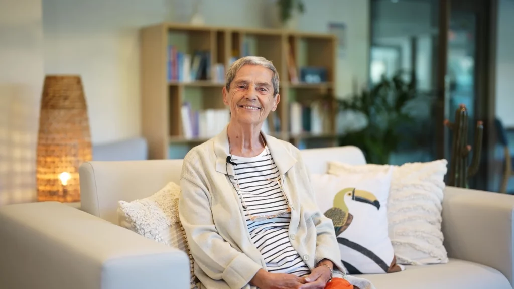 Une résidente souriante, assise sur un canapé dans un espace commun lumineux de la résidence, devant une bibliothèque