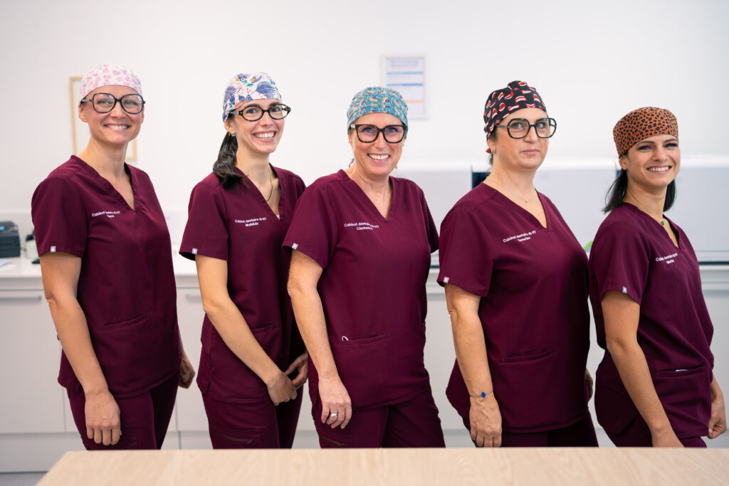 Cinq membres de l'équipe en tenue bordeaux et calots colorés, alignées et souriantes derrière un comptoir du cabinet