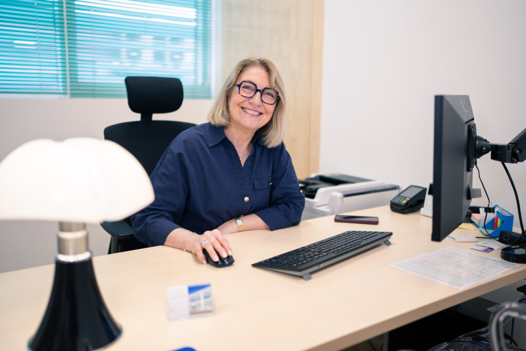 Une femme souriante en chemise bleu marine, assise à son bureau devant l'écran d'ordinateur, avec une lampe à côté d'elle