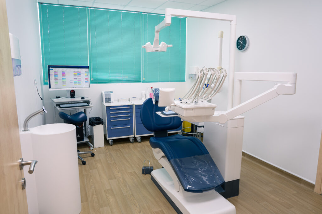 Salle de soins lumineuse avec un fauteuil dentaire à assise bleu marine, une lampe d'examen, un écran et des stores turquoise